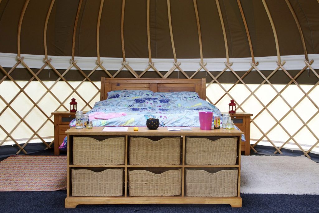 A cozy bed inside a yurt, surrounded by fabric walls and natural light filtering through a circular skylight.